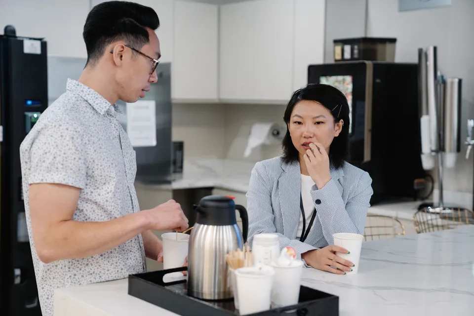 Two colleagues discussing over coffee in a modern office break room setting.